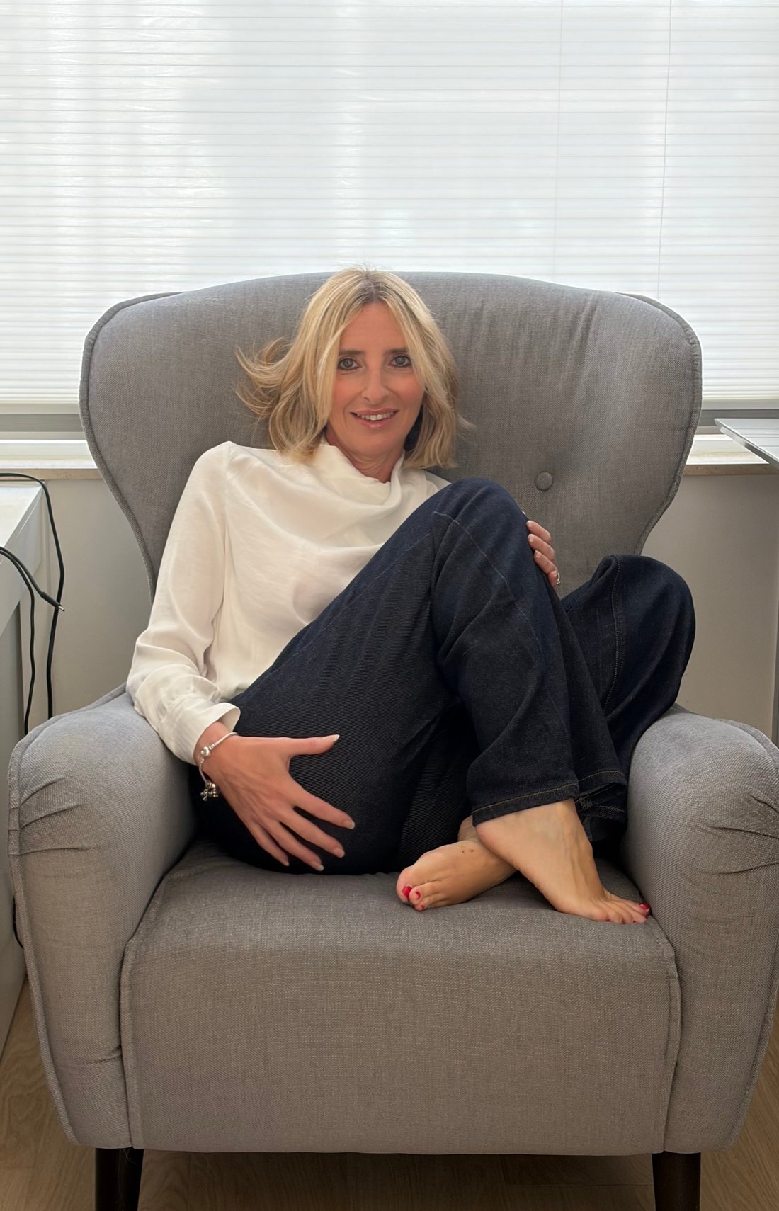 Susan Hannam curled up barefoot in a grey armchair, smiling at the camera